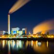 Colorful night shot of an illuminated modern industrial building with steam and deep blue sky behind a canal.