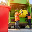 Two garbage men working together on emptying dustbins for trash removal with truck loading waste and trash bin.