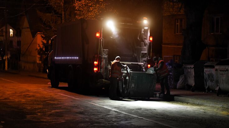 © Perytskyy - stock.adobe.com Garbage collecting truck is parked on city street. Driver prepares garbage container for loading into truck. Dark time of day.