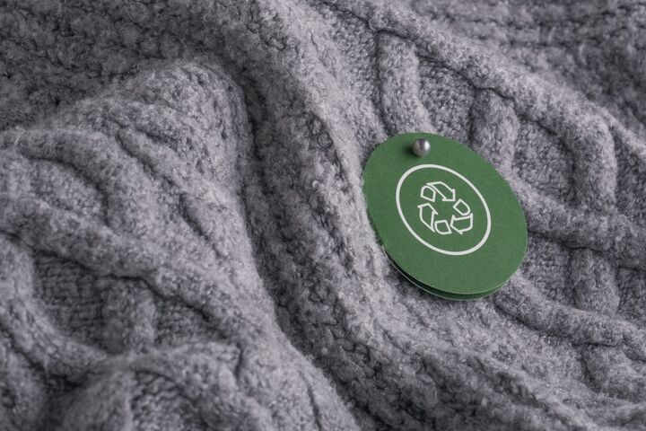 A circular green tag with a recycling symbol hangs from on gray knitted fabric. Concept of sustainable development, textile recycling, and conscious consumption in fashion. High quality photo