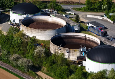 This high-resolution aerial photograph provides an excellent overview of a modern biogas and organic waste treatment facility. The image clearly shows several large circular tanks, including prominent white and black dome-covered digesters as well as open fermentation tanks. The plant is surrounded by lush green trees, agricultural fields in varying stages of cultivation, and a railway line running alongside, highlighting its integration into the rural environment.